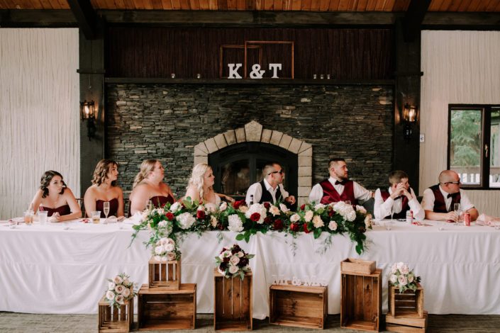 Burgundy and blush head table decorated with flower arrangements in front of a stone fireplace