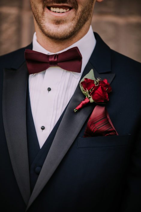 Groom boutonniere designed with a small cluster of burgundy spray roses accented by seeded eucalyptus and wrapped with burgundy satin.
