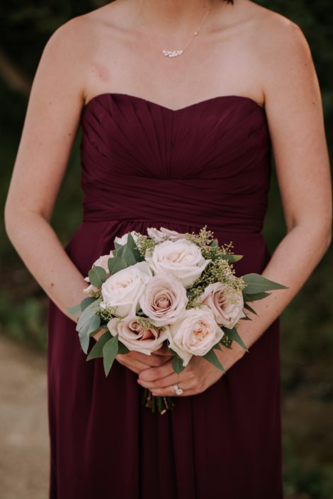 Bridesmaid wearing burgundy dress and holding blush bouquet designed with white ohara garden roses, quicksand roses and eucalyptus.