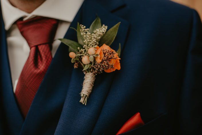 Rustic fall wedding boutonniere made of orange spray rose with hypericum, chocolate cosmos, solidago and seeded eucalyptus.