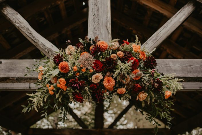 Rustic Fall Wedding archway arrangement designed with burgundy dahlias, hearts garden roses, golden mustard roses, orange spray roses, solidago, salal, italian ruscus and seeded eucalyptus.