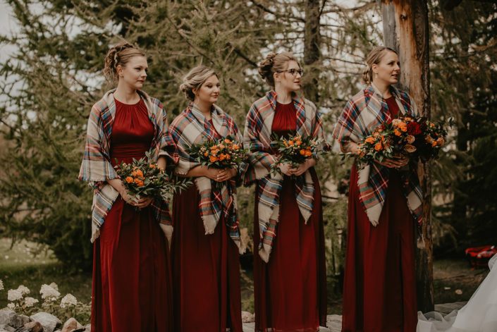 Hayley's rustic fall wedding bridesmaids wearing red dresses and plaid shawls and holding bouquets designed with orange spray roses, hypericum berries, italian ruscus and seeded eucalyptus.