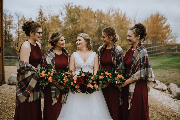 Bride and bridesmaids wearing plaid shawls for rustic fall wedding; holding bouquets designed with orange, red and yellow flowers with greenery.