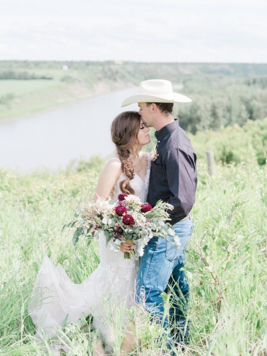 Cowboy groom forehead kissing bride; holding burgundy country chic bridal bouquet featuring burgundy dahlias, quicksand roses, olive branches, dusty miller and eucalyptus.