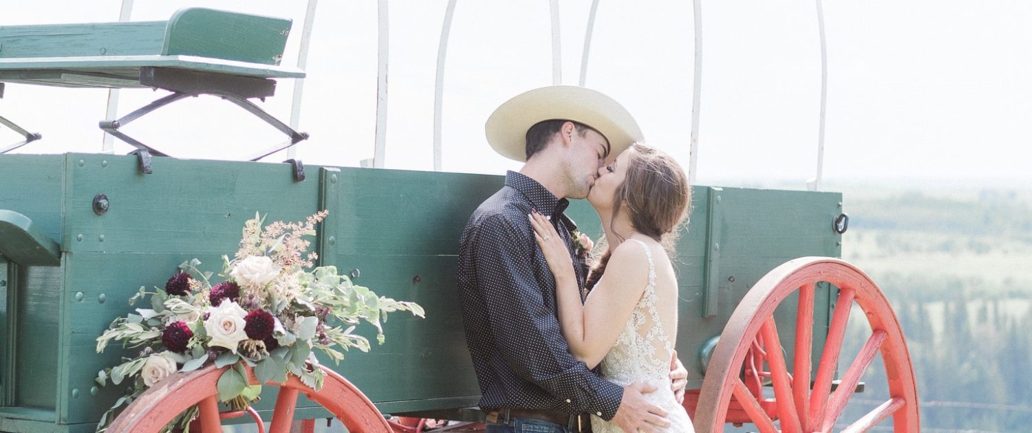 Bride and cowboy groom kissing next to a chuckwagon with a burgundy country chic bridal bouquet on the wheel.