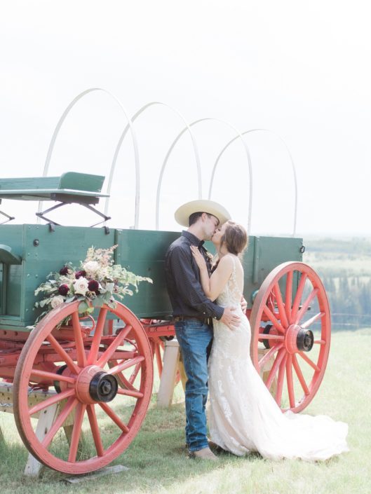 Bride and cowboy groom kissing next to a chuckwagon with a burgundy country chic bridal bouquet on the wheel.