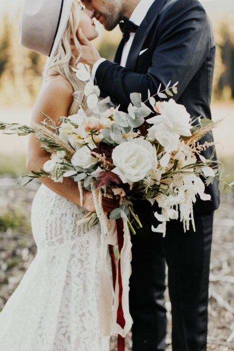 Boho bridal bouquet designed with white roses, white ranunculus, white amaranth, sweet peas, pampas grass, olive branches, eucalyptus and pops of burgundy with trailing ribbons
