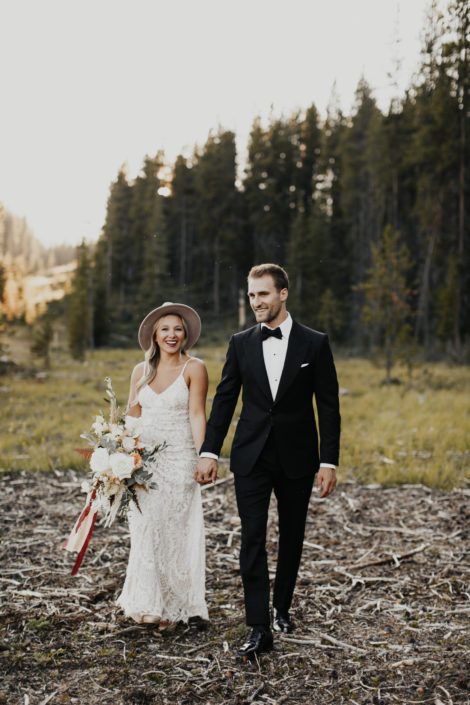 Boho bride wearing hat and carrying white bridal bouquet with burgundy accents; groom wearing tux