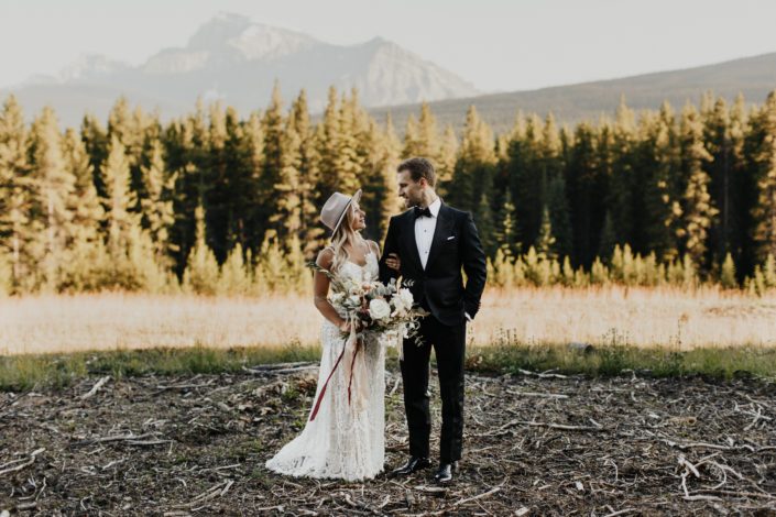 Rocky Mountain Elopement Styled Shoot - boho bride wearing lace gown, hat and carrying boho bouquet with trailing ribbons; groom wearing tux