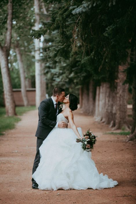 Bride and groom kissing under trees with burgundy and mauve bridal bouquet