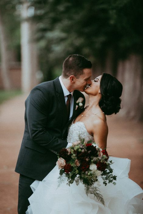 Bride and groom, Rikki Lee and Jake kissing with burgundy and mauve bridal bouquet featuring garden roses, dahlias and roses