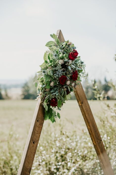Burgundy and royal blue flower arrangement atop a wooden triangle archway designed with black bacarra roses, ranunculus, eryngium, astrantia, blue viburnum berries and salal and eucalyptus greenery
