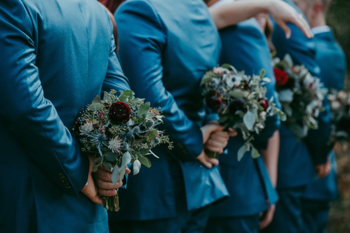 Royal blue suited groomsmen holding burgundy and blue bouquets behind their backs