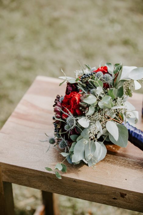 Burgundy and blue bouquet designed with black bacarra roses, ranunculus, astrantia, eryngium and blue viburnum berries wrapped with a blue satin handle