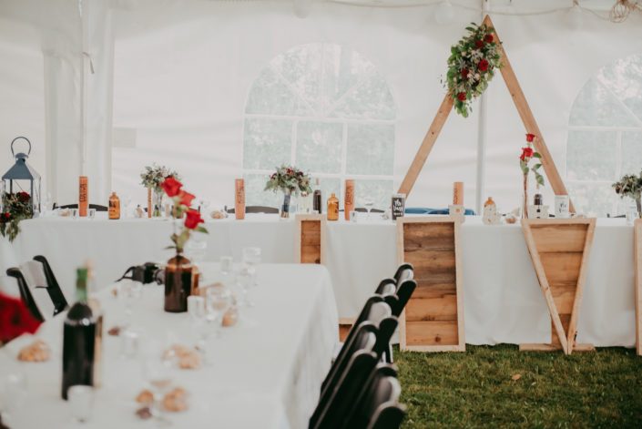 Reception for burgundy and royal blue wedding in a tent; head table with triangle archway flower arrangement and black bacarra red roses in brown bottles