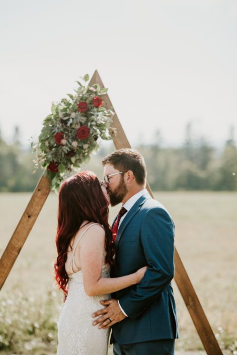 Bride and groom kissing at wedding ceremony in front of wooden triangle archway arrangement made of black bacarra roses and burgundy ranunculus