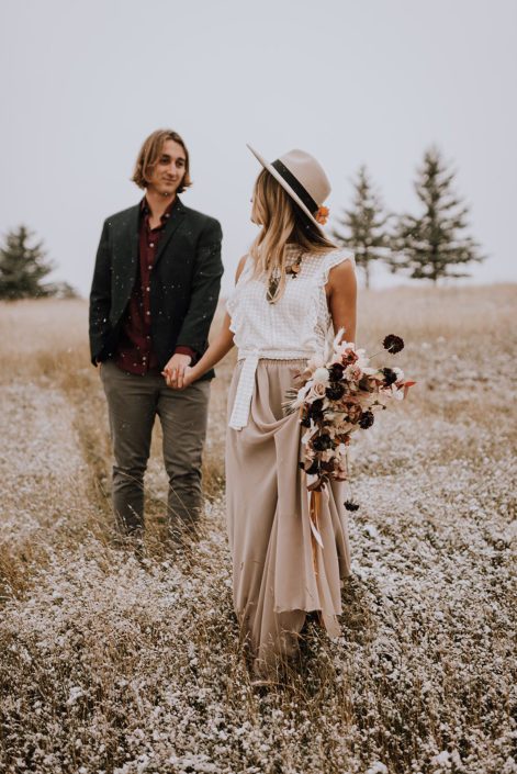 Couple walking through a field with falling snow and a neutral fall bouquet designed with plum scabiosa, roses, sweet peas, bunny tails and fall foliage