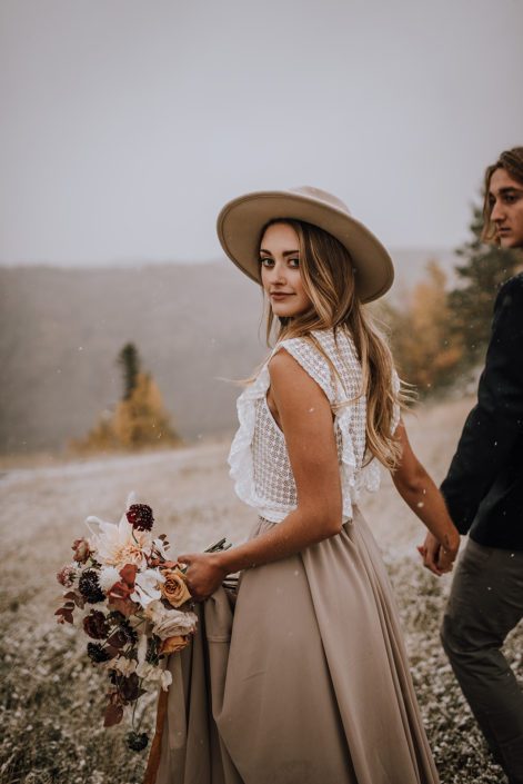 couple walking with a neutral coloured fall bouquet through a field with snow