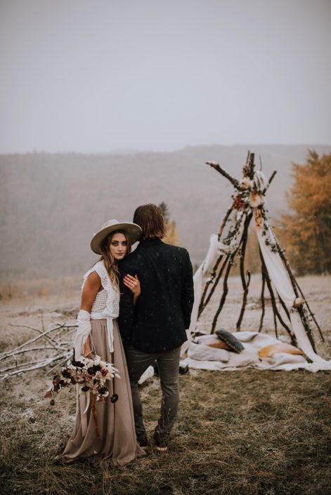 couple standing next to a tee pee for down the aisle fall styled shoot with fall coloured flowers