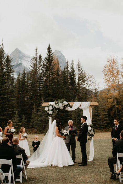Brittany and Briggs' Elegant Canmore Wedding outdoor ceremony featuring a wooden archway draped with white linens with a corner flower arrangement designed with white hydrangeas.