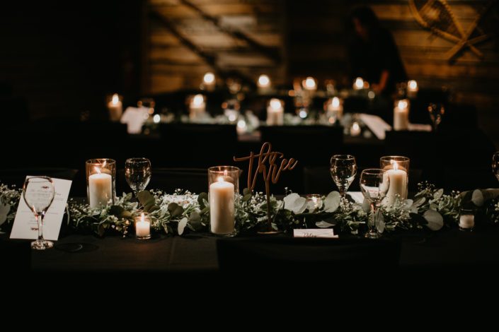 Reception decorated with long tables with black table cloths, eucalyptus runner with babies breath, candles and rose gold table number signs