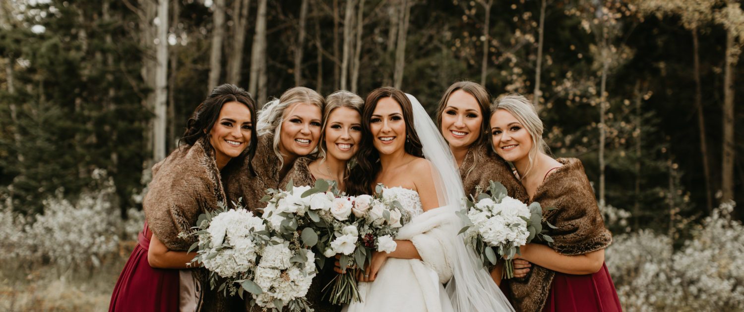 Brittany and her bridesmaids; elegant Canmore Wedding; bridesmaids wearing burgundy dresses and brown fur shawls carrying white hydrangea bouquets; bride wearing white gown and veil with white fur shawl and carrying blush and ivory bouquet featuring quicksand roses, white o'hara garden roses, lisianthus, ranunculus, astrantia and eucalyptus