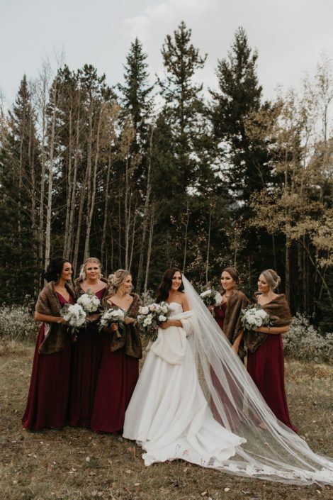 Brittany and her bridesmaids wearing fur shawls and holding ivory bouquets; bridesmaids wearing burgundy and brown shawls holding white hydrangea bouquets; bride wearing white gown and white fur shawl holding white and blush bouquet