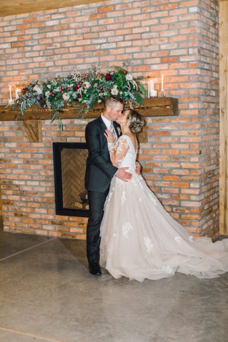 Bride and groom kissing in front of a fireplace with a blush and burgundy floral garland on the mantel