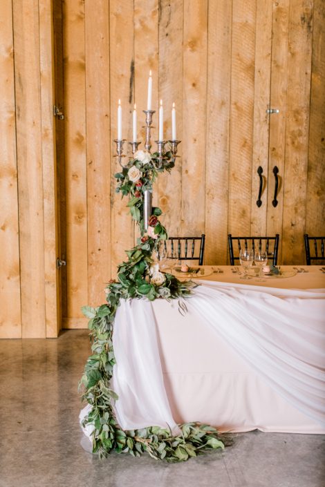 Head table tall candlestick holder decor with fresh greenery garland accented by blush and burgundy florals