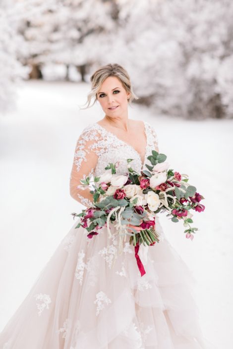 Winter bride standing in a wonderland of snow with blush and burgundy bridal bouquet