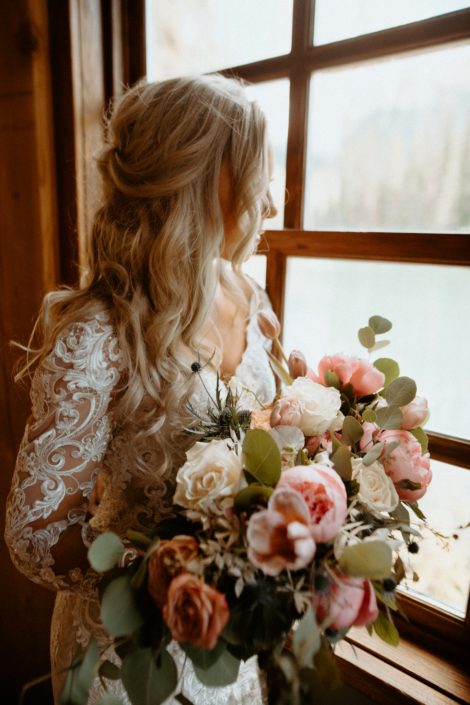 Bride looking out window with blush pink, white and blue bouquet designed with roses, tulips, peonies, eryngium and eucalyptus