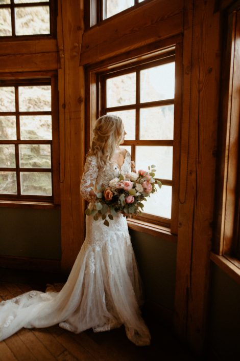 Bride standing next to window in Emerald Lake Lodge with bouquet of peonies, roses, tulips and eryngium