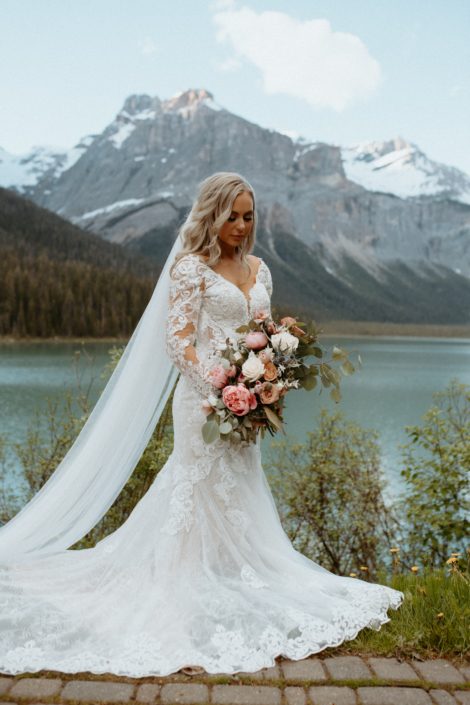 Bride holding pink and white bridal bouquet designed with peonies, roses, eryngium and tulips with a view of Emerald Lake and the Rocky Mountains
