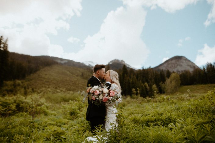 Bride and groom kissing on a fern covered mountainside with a pink and white bouquet designed with peonies, roses, and tulips