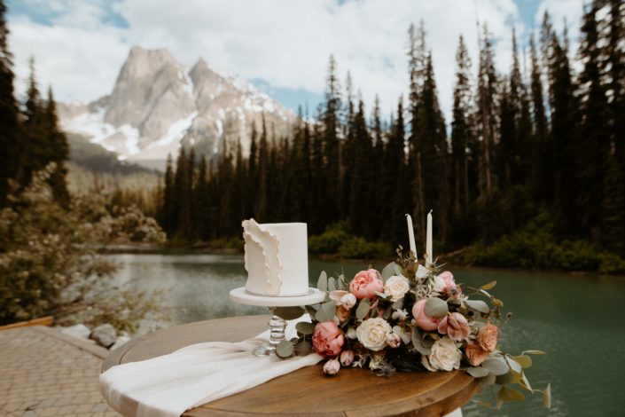 Cake Table with a view of Emerald Lake and the Rocky mountains; white ruffled wedding cake and blush pink and white bridal bouquet designed with roses, peonies and tulips