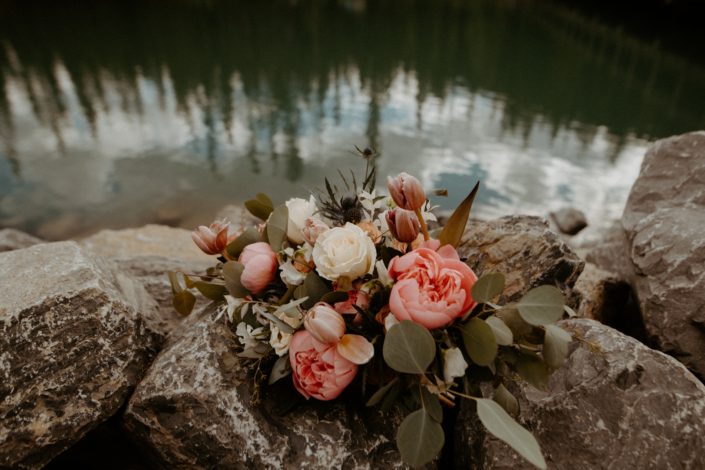 Bridal bouquet on riverside rocks featuring pink peonies, white roses, toffee roses, tulips and blue eryngium with eucalyptus greenery