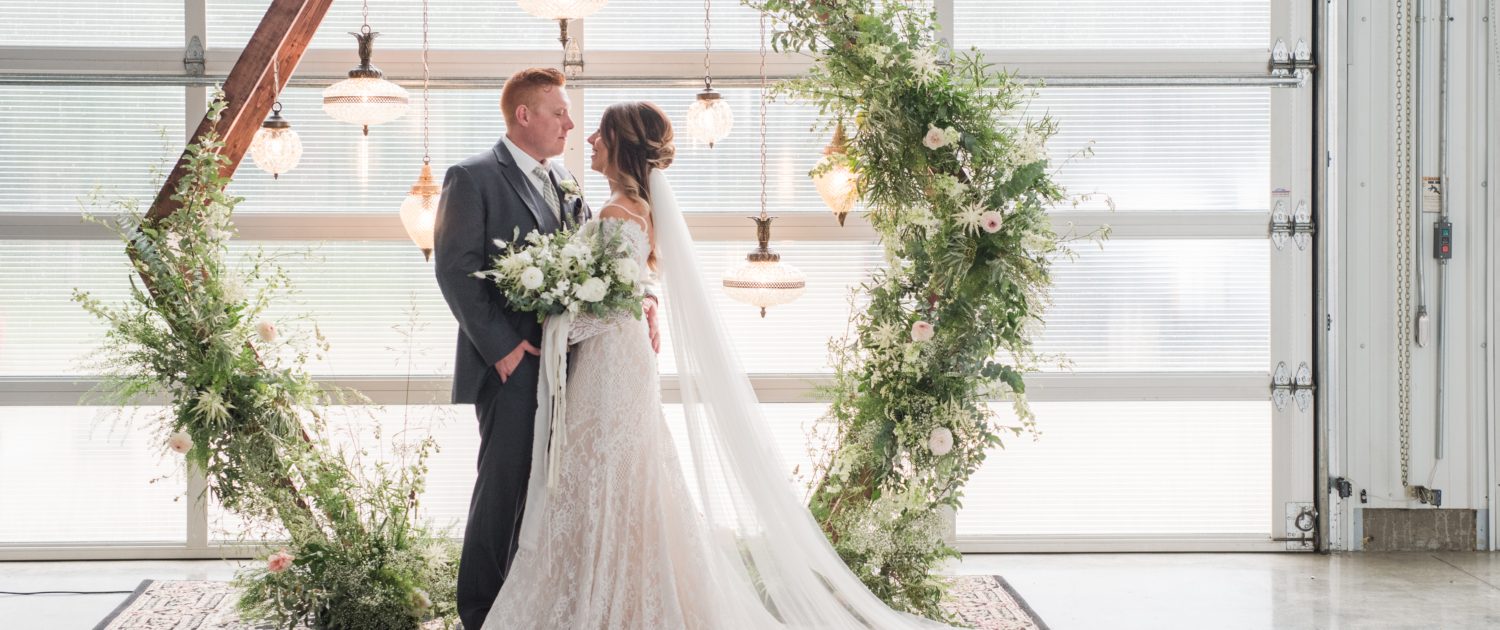 Vintage and modern, romantic industrial styled wedding shoot featuring white and blush flowers and lots of greenery; wooden hexagon archway covered with botanicals and vintage lamps; bride and groom with white and green bouquet