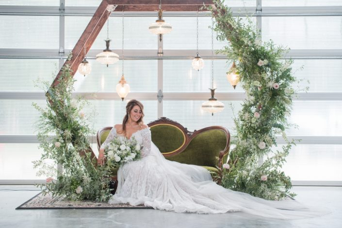 Green and white romantic industrial styled shoot - White lace gowned bride holding white and green bridal bouquet on a vintage green velvet couch under a wooden hexagon archway covered with botanicals and hanging vintage lamps.