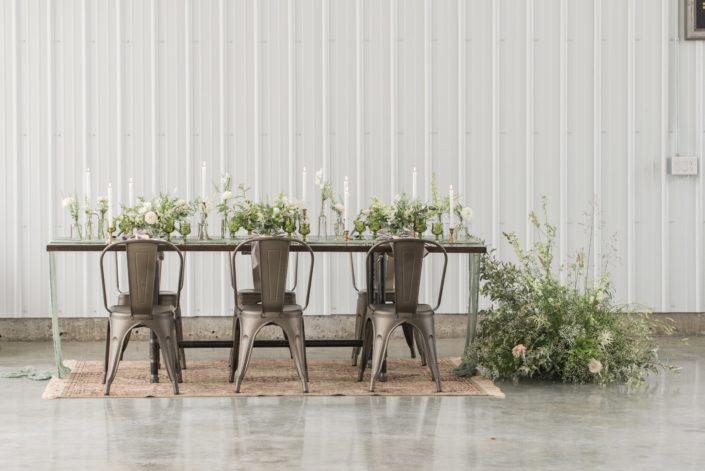 Modern, vintage, romantic industrial styled shoot table with industrial chairs, wild and natural floral arrangements designed with white flowers and lots of greenery atop a vintage persian rug.