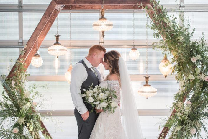 Bride and groom under wooden hexagon archway covered with green and white botanicals and hanging vintage lamps with white and green bridal bouquet featuring poppies, ranunculus, bunny tail, snapdragon, veronica and astilbe