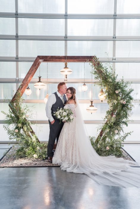 Groom kissing bride under wooden hexagon archway covered with white and green botanicals and hanging vintage lamps with white bridal bouquet
