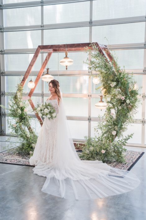 Bride holding white and green bridal bouquet beside a wooden hexagon archway covered with wild and natural green and white botanicals.