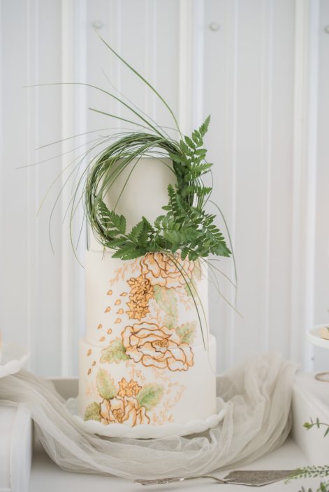 White wedding cake with gold and green accents and a wreath made of greenery (grass and leather leaf)
