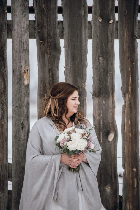 Bride wearing grey shawl and holding a mauve and ivory bouquet designed with astilbe, eryngium, white o'hara garden roses, ranunculus, amnesia roses, playa blanca roses and eucalyptus