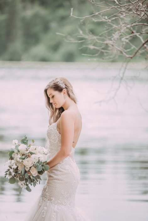 Bride standing beside a lake with a white bouquet made of roses, ranunculus, sweet peas and eucalyptus