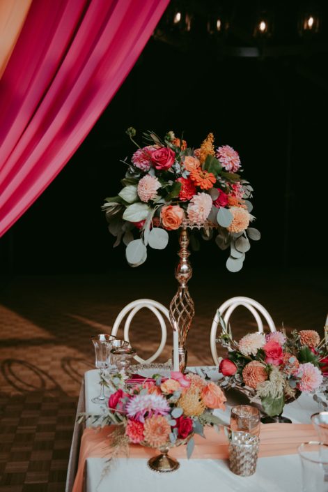 Tall arrangement featuring bold fuchsia and orange flowers such as dahlias, zinnias, and roses with eucalyptus greenery