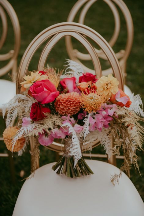 Fuchsia, orange and gold bouquet designed with zinnias, roses, dahlias, anemones, sweet peas, bleached bracken fern, and gold plumosa