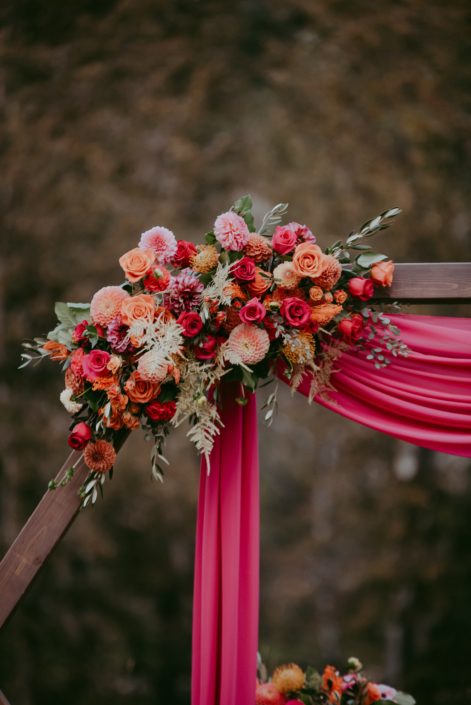 Bold fuchsia and orange wooden archway corner flower arrangement designed with roses, dahlias, zinnias, bleached bracken fern, gold plumosa and olive branches