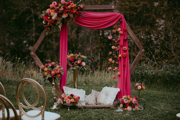 Wooden archway decorated with fuchsia drapery, and colourful fuchsia and orange flower arrangements with gold accents