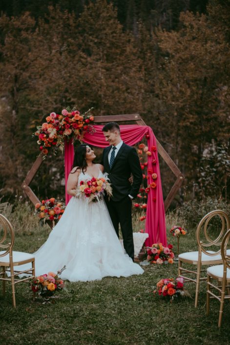Bride and groom at the end of the aisle in front of wooden hexagon archway decorated with fuchsia and orange floral arrangements and drapery